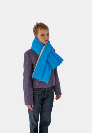 Sciarpa imbottita blu con un lato in crema. Presenta una texture morbida e una chiusura regolabile. Indossata sopra una giacca viola e jeans scuri.