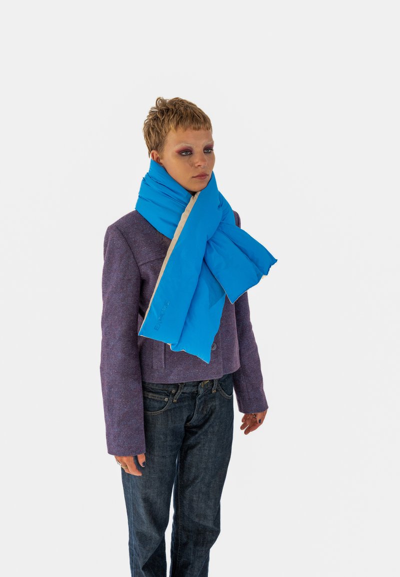 Modrá vystužená šatka s krémovou spodnou stranou. Vlastnosti zahŕňajú mäkkú textúru a nastaviteľné zapínanie. Nosená na fialovej bunde a tmavých džínsoch.