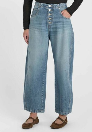 Femme portant un jean bleu taille haute à jambes larges avec cinq boutons argentés, un haut noir à manches longues, et des ballerines marron avec boucle.