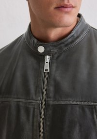 Nahaufnahme einer Person in einer dunkelgrauen Lederjacke mit silbernem Reißverschluss und Druckknopfkragen.