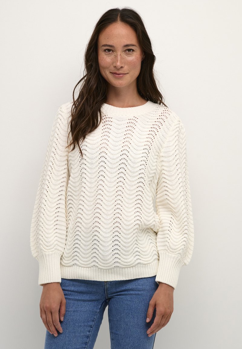 Pull en tricot blanc avec un motif en vagues, poignets et ourlet côtelés, offrant une coupe ample et un col rond, associé à un jean bleu.
