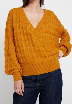 Sennepsgul strikket sweater med V-udskæring og oversized pasform. Strukturmønster i diamant, ribbede mansjetter og kant. Kombineret med mørke jeans.