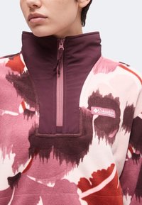 Άτομο που φοράει ροζ, κόκκινη και σκούρα μοβ fleece μπλούζα Columbia με υψηλό γιακά και μερικό φερμουάρ.