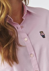 Camicia rosa chiaro in tessuto con colletto a punta, dotata di bottoni rotondi e un piccolo dettaglio ricamato di un orsetto sul lato sinistro del petto.