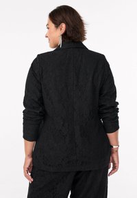 Mujer con pelo corto castaño, vestida con un blazer de encaje negro con flores y pantalones a juego, de espaldas, con pendientes colgantes y una pulsera.