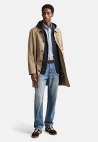 Trench beige sopra una felpa con cappuccio blu navy, camicia con bottoni e jeans azzurri. Sneakers marroni e crema completano l'outfit.