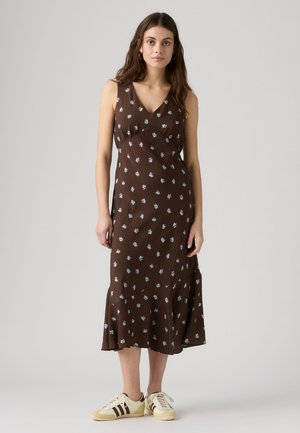 Femme aux longs cheveux bruns portant une robe marron sans manches avec de petites fleurs blanches et des baskets crème avec des rayures noires.