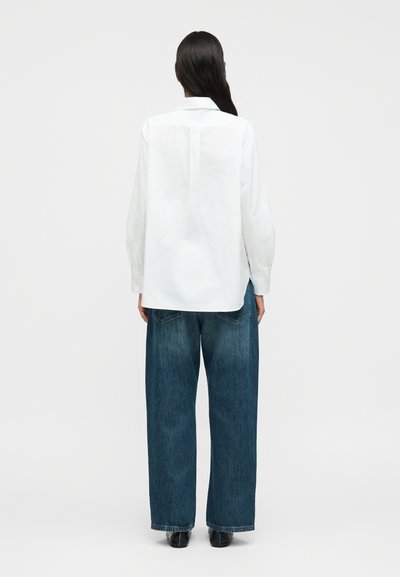 Λευκό πουκάμισο με μακριά μανίκια και κουμπωτά μανσέτα, συνδυασμένο με φαρδιά μπλε τζιν. Απλός σχεδιασμός χωρίς ορατά σχέδια ή διακοσμήσεις.