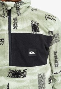 Fleece-Pullover in Hellgrün mit schwarzen Akzenten, mit abstrakten Mustern. Mit Reißverschluss vorne, Stehkragen und Logopatch auf der Brust.