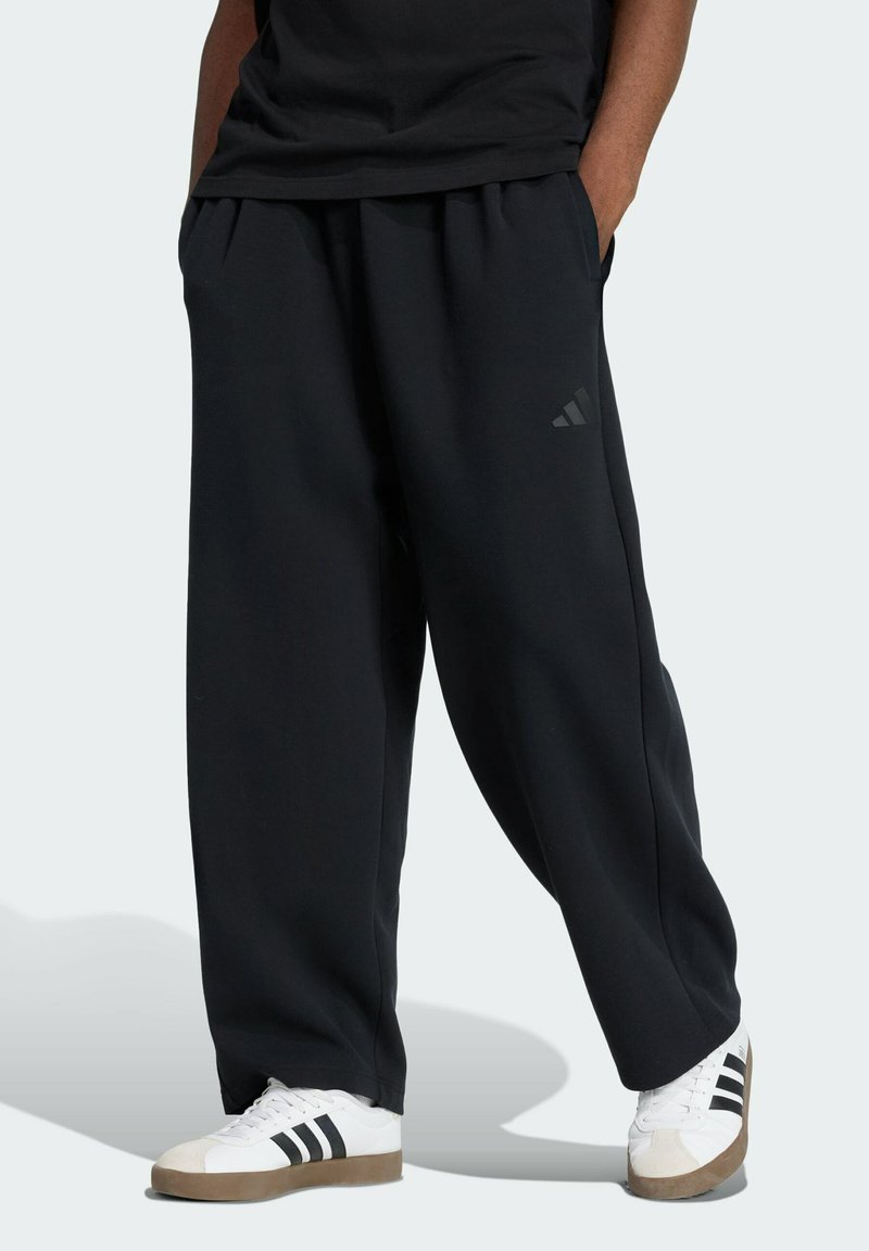 Black sweatpants with a relaxed fit, pleated front, and side pockets. Features a subtle dark logo on the leg and a soft cotton-like texture.