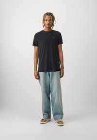 Schwarzes T-Shirt mit kurzen Ärmeln und Logo auf der Brust, kombiniert mit hellblauen, weiten Jeans und braunen Schuhen mit weißen Akzenten.