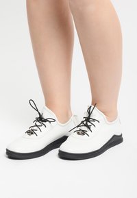 Zapatillas de malla blancas con cordones y suela negra. Punta redondeada y diseño bajo. Presenta una superficie texturizada y acentos metálicos en los cordones.