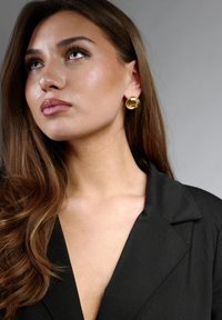 Pendientes circulares de oro con un patrón texturizado, llevados con una blazer negra de escote en V profundo y solapas afiladas, tono de piel suave.