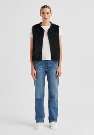 Frau in blauer Jeans, weißen Turnschuhen, weißem T-Shirt und schwarzer, ärmelloser, flauschiger Weste vor schlichtem hellem Hintergrund stehend.