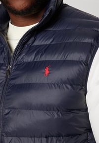 Man som bär en marinblå vadderad väst med en röd broderad polospelarlogotyp över en vit långärmad skjorta.