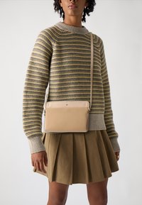 Beige Umhängetasche aus glattem Leder mit einfachem Design und subtiler Markenkennzeichnung, kombiniert mit einem gestreiften Pullover und einem plissierten Rock.