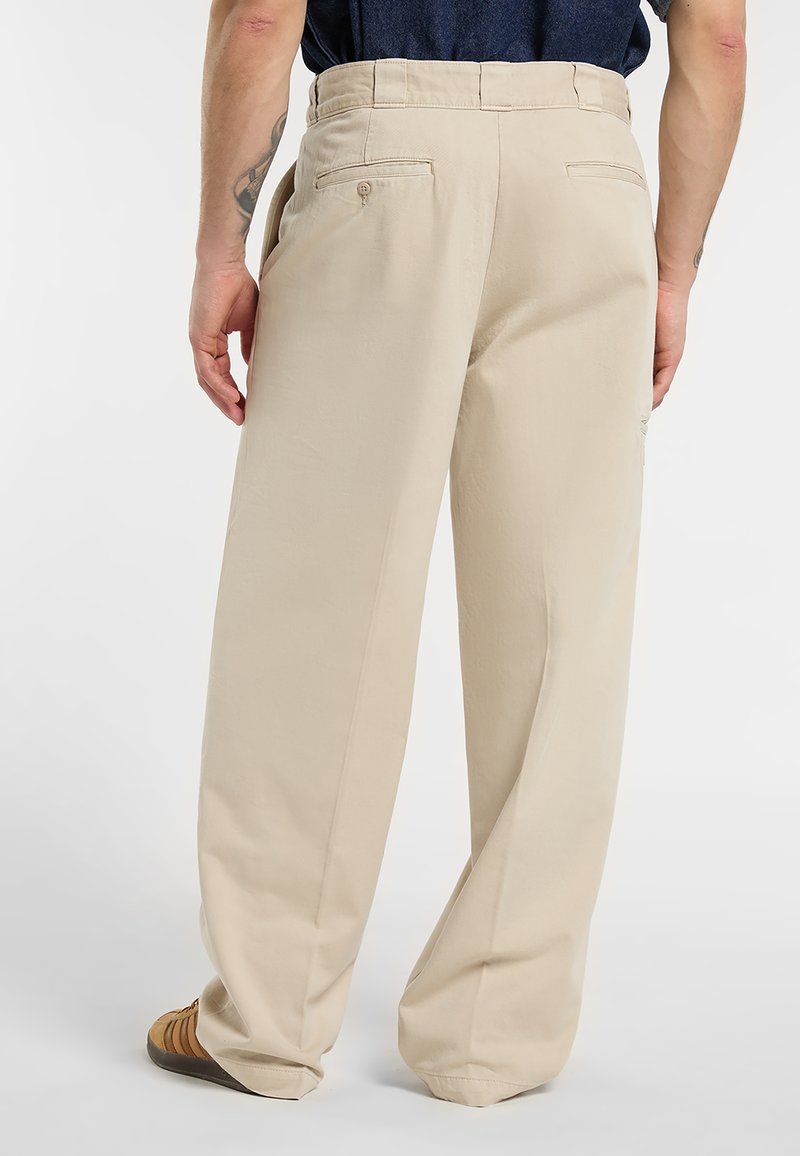 Uomo con pantaloni beige a vestibilità larga e sneaker marroni, visto da dietro su sfondo bianco semplice.