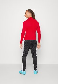 Rotes langärmliges Sporthemd, schwarze Hose mit weißen Streifen, blaue Fußballschuhe. Glatte Textur, taillierte Passform, minimale Details.