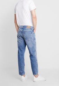 Henkilö, jolla on vaaleansiniset suorat farkut takataskuilla ja valkoinen t-paita, seisoo kädet taskuissa yksinkertaista taustaa vasten.