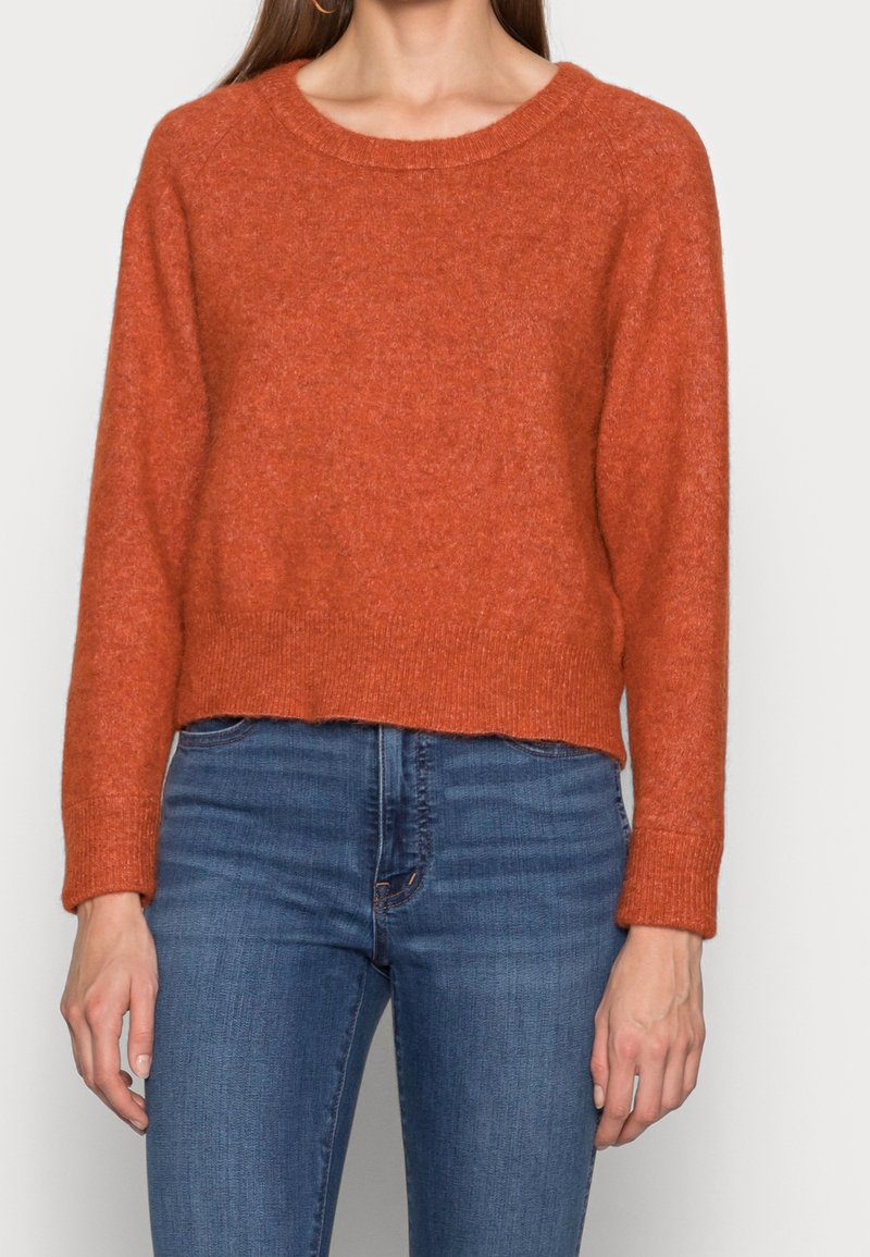 Femme portant un pull en tricot couleur rouille à manches longues et un jean bleu taille haute sur un fond neutre.
