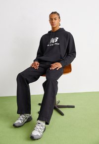 Fekete kapucnis pulóver "new balance" logóval, fekete farmernadrág és szürke futócipő. Egy fa széken ül, fehér háttér előtt. Zöld padló.