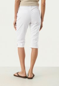 Pantalon capri blanc à coupe slim, avec une taille mi-haute, des poches arrière et de légères fentes sur l'ourlet. Porté avec des sandales marron.