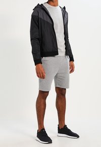 Černá lehká bunda na zip s kapucí, šedé tričko s kulatým výstřihem a šedé šortky, doplněné černými sportovními botami.