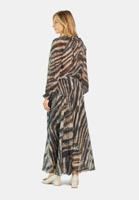 Maxikleid aus transparenter, gemusterter Stoff mit dunklen und hellen Wellenstreifen. Es hat einen gerafften Ausschnitt und lange Ärmel, kombiniert mit Blockabsatzschuhen.