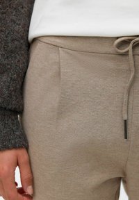 Béžové kalhoty z měkké strukturované tkaniny, s vytvořeným pasem na šňůrku a bočními kapsami, doplněné tmavým pleteným svetrem.