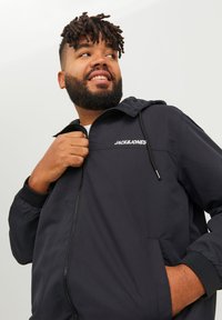 Black zip-up jacket with a hood, featuring a white logo on the chest, elastic cuffs, and a smooth, lightweight texture.