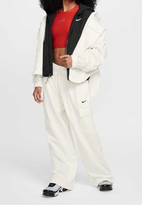 Bílé cargo kalhoty, zkrácený červený top a černobílá vrstvená bunda. Mezi rysy patří zipové uzávěry a akcenty s logem Nike.