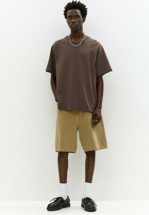 Brun oversized kortærmet sweatshirt, beige knæ-lange shorts, sorte sko og hvide sokker. Accessories inkluderer lagdelte sølv halskæder.