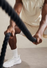Scarpe da ginnastica bianche con dettagli in rete e calze a coste. Una persona in una canottiera chiara e pantaloncini afferra una corda spessa nera.