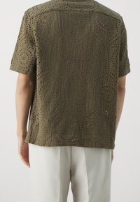 Olijfgroen shirt met korte mouwen, versierd met ingewikkelde uitsnijdpatronen en een textuurafwerking. Het heeft een ontspannen pasvorm en een rechte zoom.