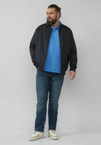 Marineblauer Bomberjacke über einem hellblauen Poloshirt, kombiniert mit blauen Jeans und weißen Turnschuhen. Die Jacke hat einen Reißverschluss und seitliche Taschen.