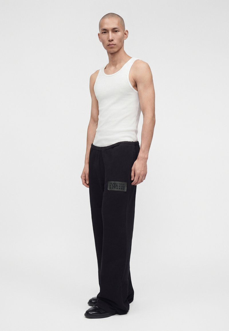 Homme debout portant un débardeur blanc sans manches, un pantalon noir ample avec un patch et des chaussures noires sur un fond clair uni.