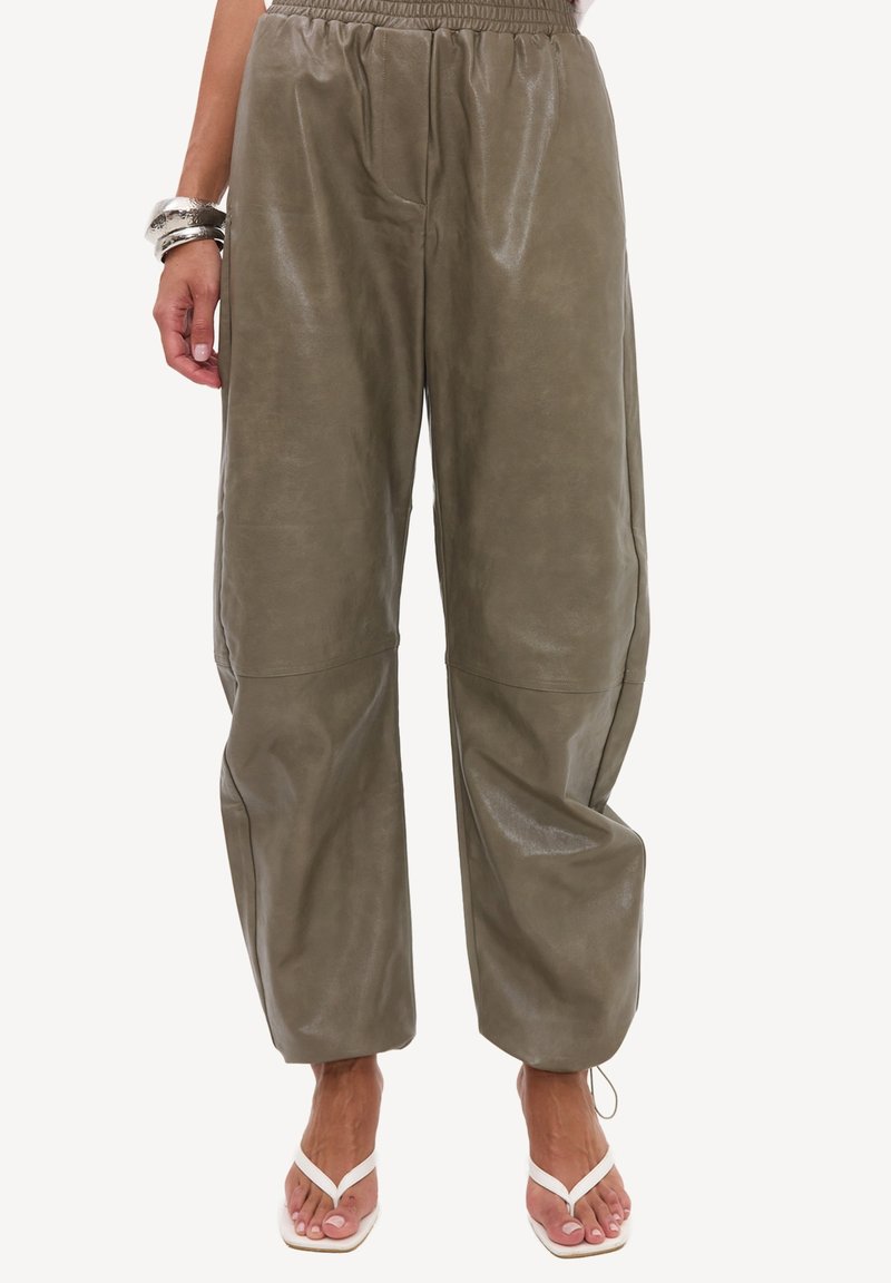 Pantalon ample vert olive avec taille élastique et coutures au-dessus des genoux, porté avec des talons blancs à brides et des bracelets argentés au poignet gauche.