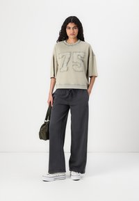 Ljusgrön kort sweatshirt med stort broderat "75" på framsidan, tillsammans med löst sittande mörkgrå byxor och vita sneakers. Olivgrön väska över axeln.