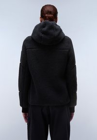 Chaqueta de forro polar negra con capucha, superficie texturizada y hombros caídos. Diseño simple, con un corte holgado y sin cierres visibles.