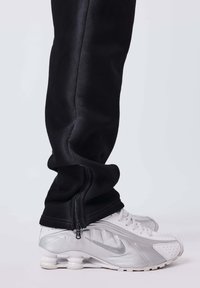 Piernas vestidas con pantalones negros con cremallera lateral sobre zapatillas Nike blancas que presentan un exclusivo diseño de suela en forma de resorte.