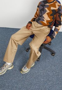 Koszula z długim rękawem w zygzakowy wzór w kolorze brązowym, czarnym i szarym; beżowe spodnie; szare sneakersy z żółtymi akcentami; siedzący na niebieskim krześle.