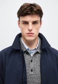 Νεαρός άνδρας με κοντά καστανά μαλλιά, φοράει μπλε μπουφάν πάνω από γκρι πουλόβερ με κουμπιά και μπλε-λευκό ριγέ πουκάμισο, κοιτάζοντας μπροστά.