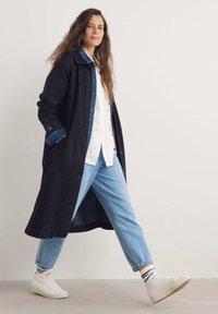 Lång svart denimjacka med en kontrasterande blå krage, knäppning framtill och sidofickor, kombinerad med ljusblå jeans och vita sneakers.