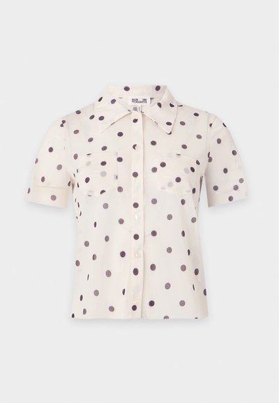 Blouse crème à manches courtes avec des pois noirs, boutons à l’avant, deux poches poitrine et un large col pointu.