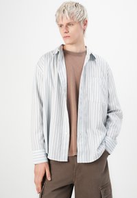 Light blue striped shirt with brown vertical stripes, long sleeves, and a single chest pocket, worn over a brown t-shirt and brown pants.