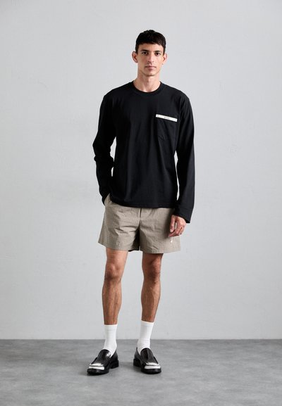 Zwarte longsleeve met een borstzak, gecombineerd met lichtbruine geruite shorts. Zwarte schoenen met witte accenten en witte sokken.