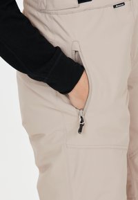 Lichtbeige broek met een ritszak, gemaakt van stevig materiaal. Een zwart shirt met lange mouwen is deels zichtbaar bij de mouw.