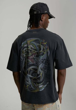 Hombre con el cabello trenzado que lleva una gorra negra y una camiseta negra con un diseño colorido de serpiente y pájaros junto con caracteres asiáticos en la parte trasera.