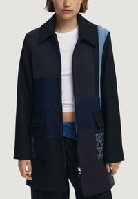 Cappotto patchwork nero con un mix di texture, tra cui denim e tessuto a coste. Presenta un colletto, chiusura con zip e due tasche frontali.