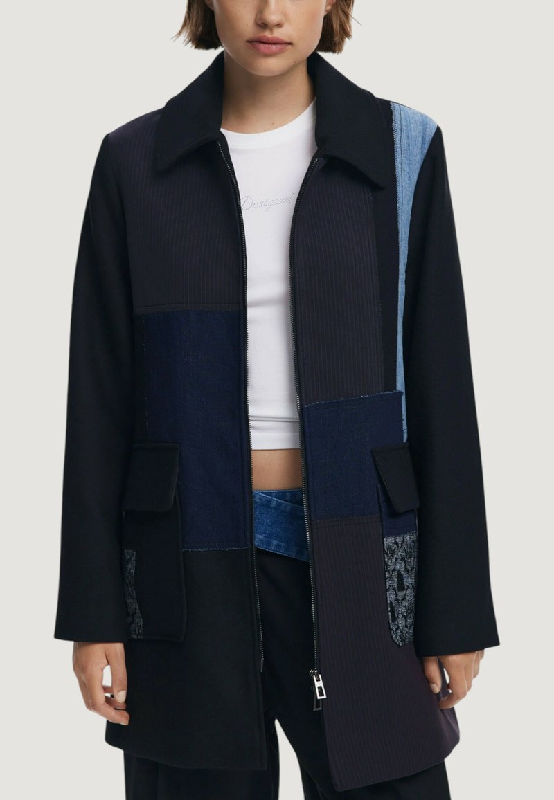 Cappotto patchwork nero con un mix di texture, tra cui denim e tessuto a coste. Presenta un colletto, chiusura con zip e due tasche frontali.